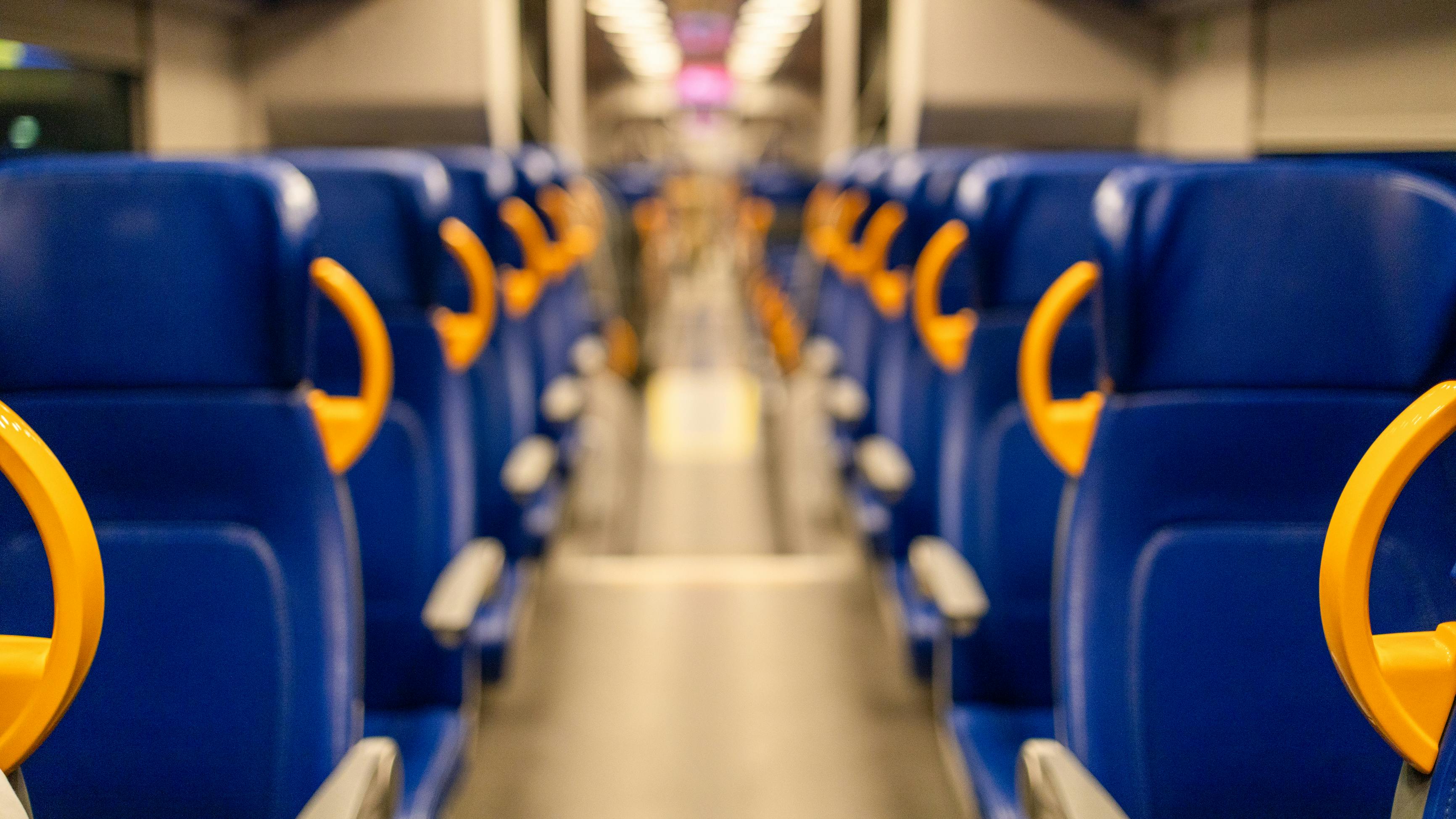 A shallow-depth-of-field photograph looking down the center aisle of an empty passenger rail car. Rows of blue upholstered seats with bright yellow/orange plastic grab handles line both sides. Overhead luggage racks and fluorescent ceiling lights recede toward a blurred doorway at the far end. The floor is light gray and the car appears to be at rest.