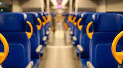 A shallow-depth-of-field photograph looking down the center aisle of an empty passenger rail car. Rows of blue upholstered seats with bright yellow/orange plastic grab handles line both sides. Overhead luggage racks and fluorescent ceiling lights recede toward a blurred doorway at the far end. The floor is light gray and the car appears to be at rest.