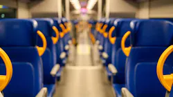 A shallow-depth-of-field photograph looking down the center aisle of an empty passenger rail car. Rows of blue upholstered seats with bright yellow/orange plastic grab handles line both sides. Overhead luggage racks and fluorescent ceiling lights recede toward a blurred doorway at the far end. The floor is light gray and the car appears to be at rest. A shallow-depth-of-field photograph looking down the center aisle of an empty passenger rail car. Rows of blue upholstered seats with bright yellow/orange plastic grab handles line both sides. Overhead luggage racks and fluorescent ceiling lights recede toward a blurred doorway at the far end. The floor is light gray and the car appears to be at rest.