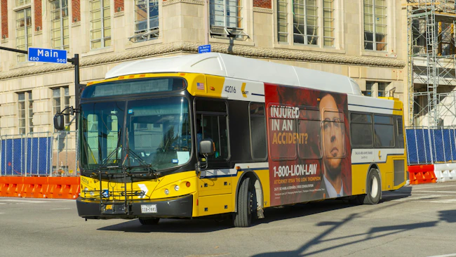 A yellow and white Dallas Area Rapid Transit bus, fleet number 42016, travels through a downtown Dallas intersection at the 400-500 block of Main Street. The bus carries a full-side advertisement for a personal injury law firm. Orange construction barriers and chain-link fencing line the street, and a multi-story building is visible in the background.