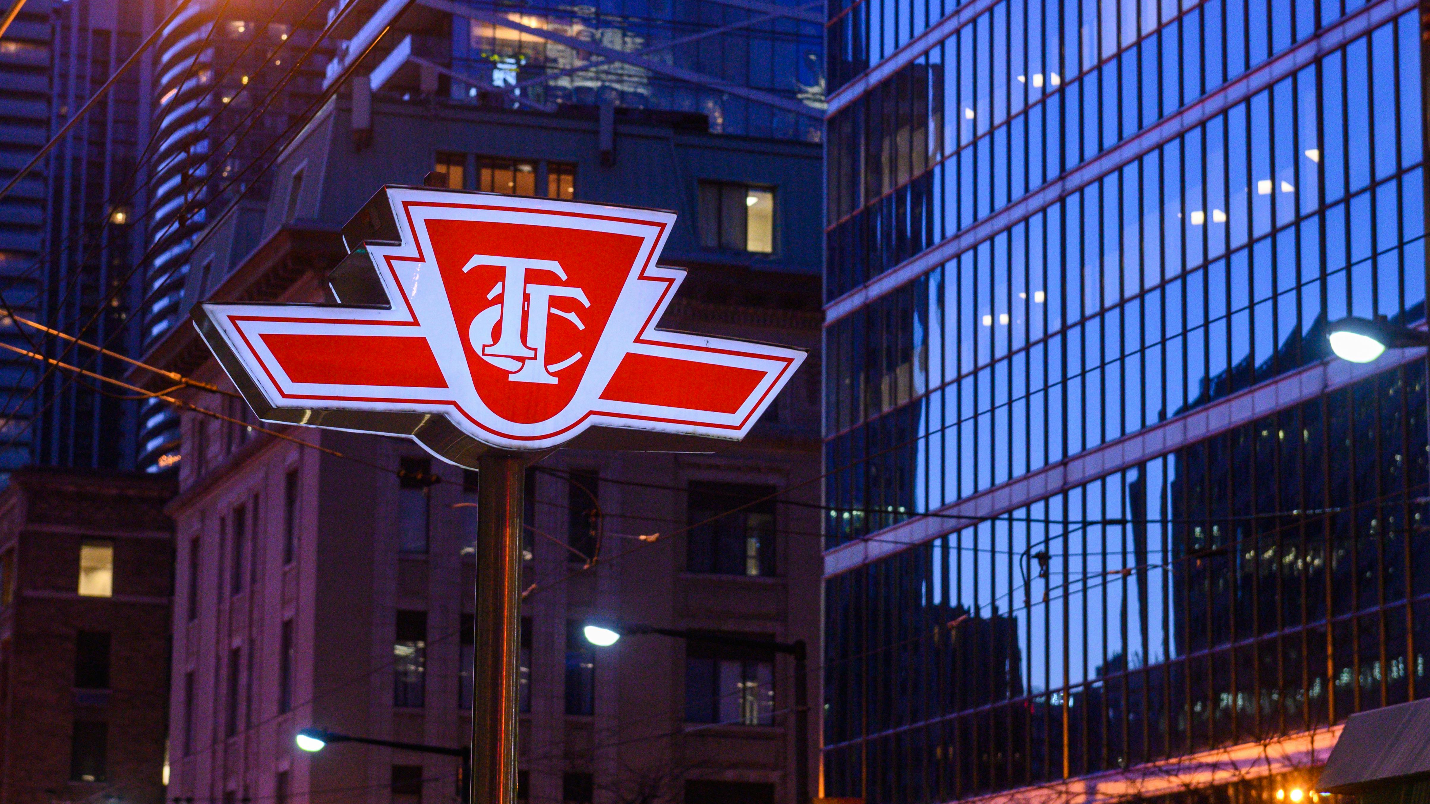 A pole-mounted Toronto Transit Commission (TTC) logo sign is photographed at dusk in downtown Toronto. The red-and-white emblem &mdash; a stylized shield with interlocking 'T' and 'C' letters flanked by horizontal speed lines &mdash; is illuminated from behind, making it glow against a blue-toned urban backdrop. Behind the sign, a mix of older brick buildings and modern glass-curtain-wall towers line the street, their windows lit from within. Overhead wires cross the frame, suggesting a streetcar corridor.