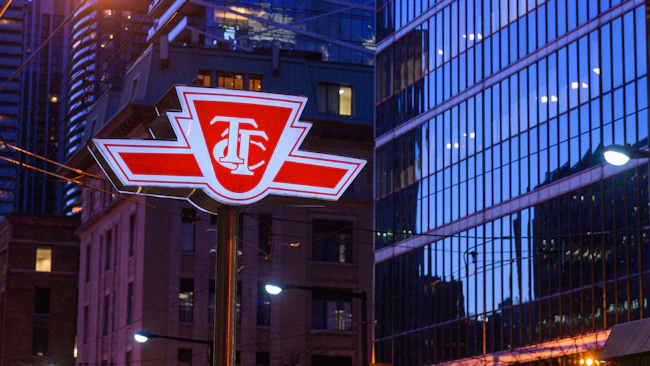 A pole-mounted Toronto Transit Commission (TTC) logo sign is photographed at dusk in downtown Toronto. The red-and-white emblem &mdash; a stylized shield with interlocking 'T' and 'C' letters flanked by horizontal speed lines &mdash; is illuminated from behind, making it glow against a blue-toned urban backdrop. Behind the sign, a mix of older brick buildings and modern glass-curtain-wall towers line the street, their windows lit from within. Overhead wires cross the frame, suggesting a streetcar corridor.