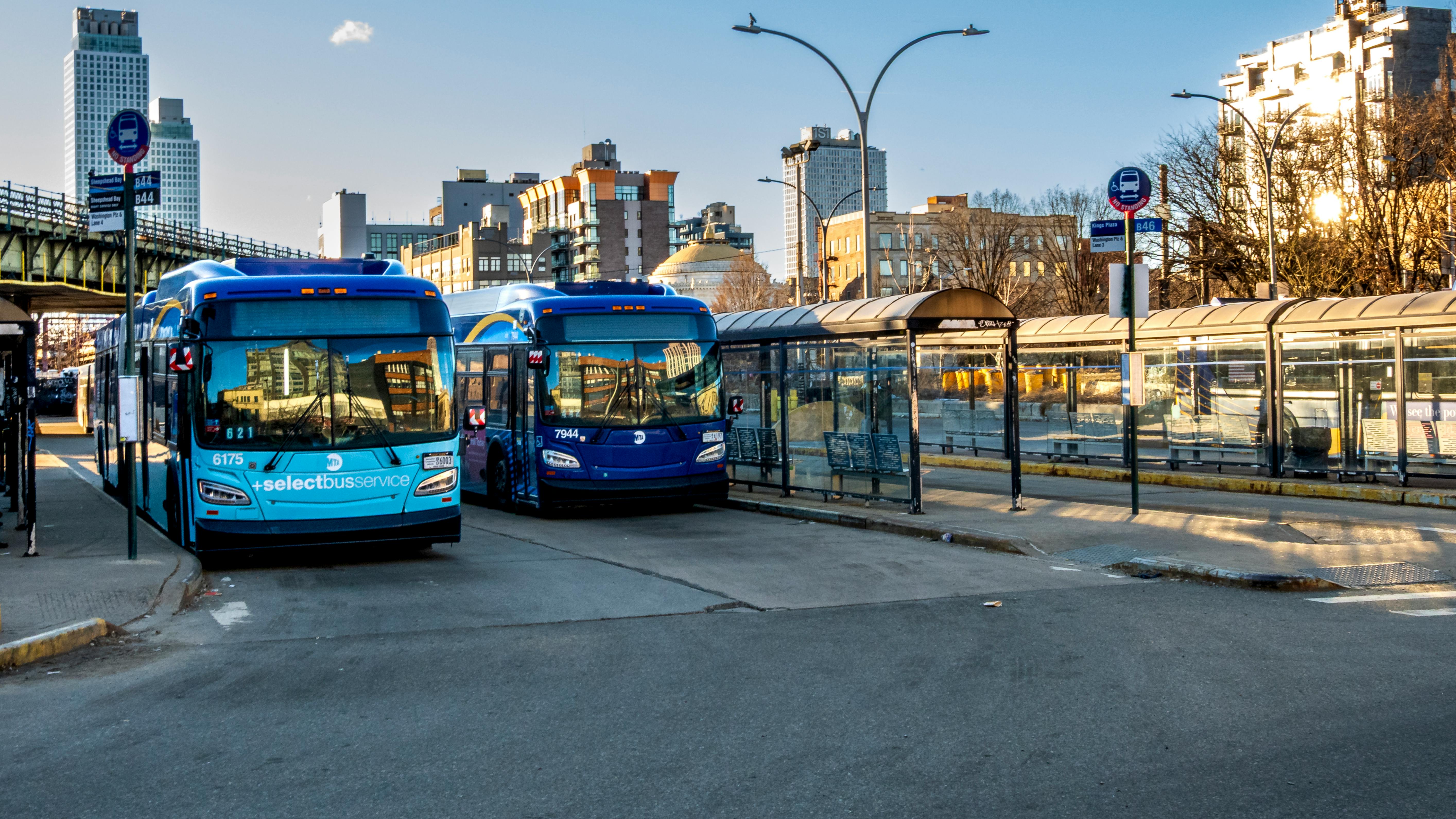 Two MTA New York City Transit articulated buses &mdash; one marked 'Select Bus Service' (fleet number 6175) and one in standard blue livery (fleet number 7944) &mdash; are stopped at a bus terminal with glass-enclosed shelters and route signage. An elevated rail structure is visible to the left, and a city skyline rises in the background under a clear blue sky at golden hour.