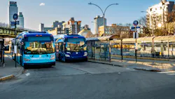 Two MTA New York City Transit articulated buses — one marked 'Select Bus Service' (fleet number 6175) and one in standard blue livery (fleet number 7944) — are stopped at a bus terminal with glass-enclosed shelters and route signage. An elevated rail structure is visible to the left, and a city skyline rises in the background under a clear blue sky at golden hour. Two MTA New York City Transit articulated buses — one marked 'Select Bus Service' (fleet number 6175) and one in standard blue livery (fleet number 7944) — are stopped at a bus terminal with glass-enclosed shelters and route signage. An elevated rail structure is visible to the left, and a city skyline rises in the background under a clear blue sky at golden hour.
