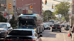 An MTA articulated bus (fleet number 6274) travels along a congested urban street in Queens, New York, surrounded by heavy car traffic. Green traffic signals are visible overhead, and a mix of commercial signage — including multilingual signs in English, Hindi and Arabic — lines the sidewalk. A large brick building with rooftop graffiti dominates the background under a partly cloudy sky. An MTA articulated bus (fleet number 6274) travels along a congested urban street in Queens, New York, surrounded by heavy car traffic. Green traffic signals are visible overhead, and a mix of commercial signage — including multilingual signs in English, Hindi and Arabic — lines the sidewalk. A large brick building with rooftop graffiti dominates the background under a partly cloudy sky.