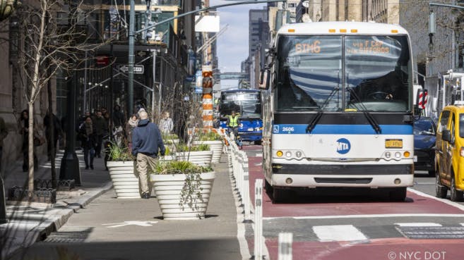 An MTA bus travels in a red-painted offset bus lane on Lexington Avenue in Manhattan, with construction equipment and workers visible in the background. Large white planters with greenery line the sidewalk to the left. A yellow taxi is partially visible on the right.