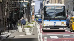 An MTA bus travels in a red-painted offset bus lane on Lexington Avenue in Manhattan, with construction equipment and workers visible in the background. Large white planters with greenery line the sidewalk to the left. A yellow taxi is partially visible on the right. An MTA bus travels in a red-painted offset bus lane on Lexington Avenue in Manhattan, with construction equipment and workers visible in the background. Large white planters with greenery line the sidewalk to the left. A yellow taxi is partially visible on the right.