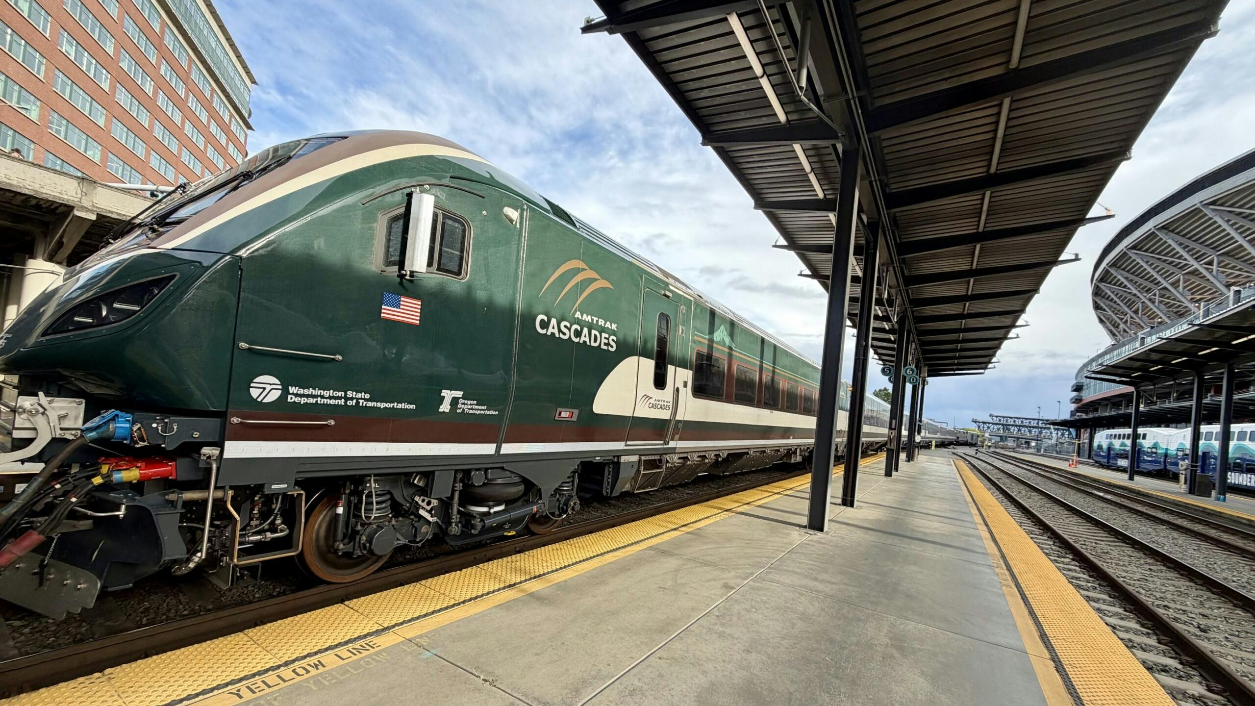 An Amtrak Cascades passenger trainset sits at a station platform, with the locomotive's dark green livery, American flag decal, and Washington State Department of Transportation and Oregon Department of Transportation logos visible on the cab nose. A covered platform with steel canopy extends to the right, with a Sound Transit Sounder commuter rail trainset visible on an adjacent track in the background.