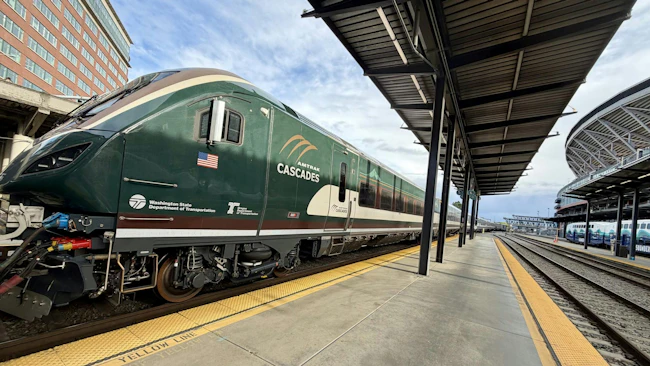 An Amtrak Cascades passenger trainset sits at a station platform, with the locomotive's dark green livery, American flag decal, and Washington State Department of Transportation and Oregon Department of Transportation logos visible on the cab nose. A covered platform with steel canopy extends to the right, with a Sound Transit Sounder commuter rail trainset visible on an adjacent track in the background.