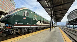 An Amtrak Cascades passenger trainset sits at a station platform, with the locomotive's dark green livery, American flag decal, and Washington State Department of Transportation and Oregon Department of Transportation logos visible on the cab nose. A covered platform with steel canopy extends to the right, with a Sound Transit Sounder commuter rail trainset visible on an adjacent track in the background.