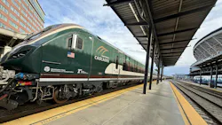 An Amtrak Cascades passenger trainset sits at a station platform, with the locomotive's dark green livery, American flag decal, and Washington State Department of Transportation and Oregon Department of Transportation logos visible on the cab nose. A covered platform with steel canopy extends to the right, with a Sound Transit Sounder commuter rail trainset visible on an adjacent track in the background. An Amtrak Cascades passenger trainset sits at a station platform, with the locomotive's dark green livery, American flag decal, and Washington State Department of Transportation and Oregon Department of Transportation logos visible on the cab nose. A covered platform with steel canopy extends to the right, with a Sound Transit Sounder commuter rail trainset visible on an adjacent track in the background.