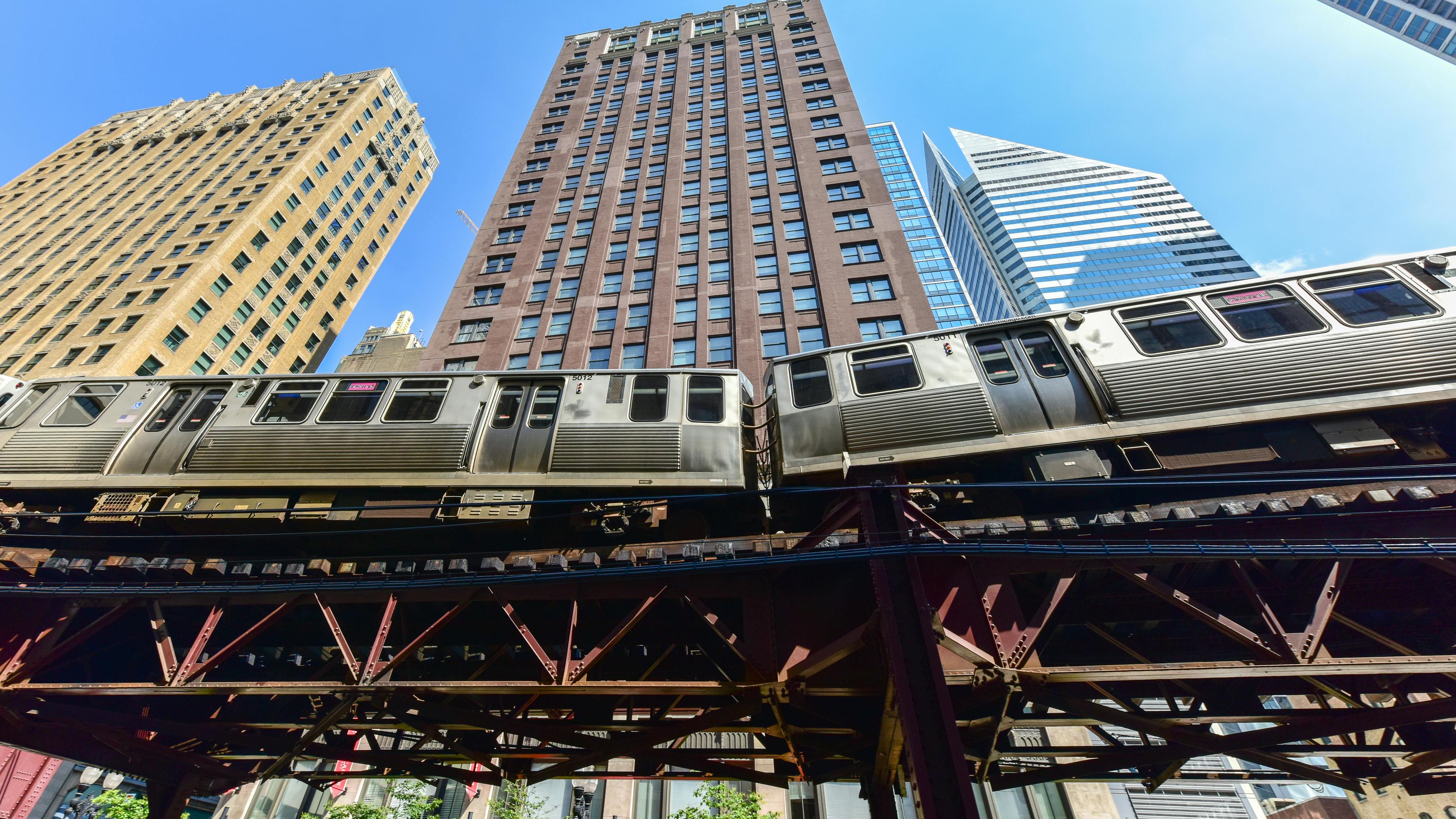 A Chicago Transit Authority elevated rail train passes through the Loop on the iconic 'L' structure, photographed from street level against a clear blue sky with downtown skyscrapers rising in the background.