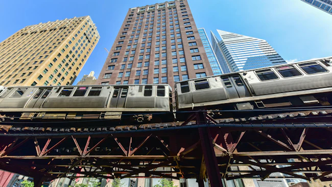 A Chicago Transit Authority elevated rail train passes through the Loop on the iconic 'L' structure, photographed from street level against a clear blue sky with downtown skyscrapers rising in the background.