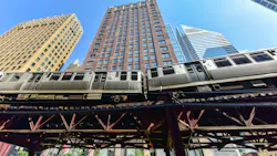 A Chicago Transit Authority elevated rail train passes through the Loop on the iconic 'L' structure, photographed from street level against a clear blue sky with downtown skyscrapers rising in the background. A Chicago Transit Authority elevated rail train passes through the Loop on the iconic 'L' structure, photographed from street level against a clear blue sky with downtown skyscrapers rising in the background.
