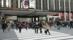 Pedestrians cross a busy midtown Manhattan intersection outside the 34th Street entrance to New York Penn Station, marked with Amtrak signage. A Chase Bank advertisement and Madison Square Garden branding are visible on the building above the entrance.