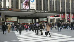 Pedestrians cross a busy midtown Manhattan intersection outside the 34th Street entrance to New York Penn Station, marked with Amtrak signage. A Chase Bank advertisement and Madison Square Garden branding are visible on the building above the entrance. Pedestrians cross a busy midtown Manhattan intersection outside the 34th Street entrance to New York Penn Station, marked with Amtrak signage. A Chase Bank advertisement and Madison Square Garden branding are visible on the building above the entrance.