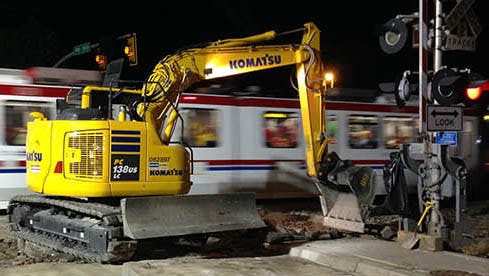 A yellow Komatsu excavator works at a grade crossing improvement site at night as a Utah Transit Authority light rail train passes in a blur of motion in the background. A railroad crossing signal is visible at right.
