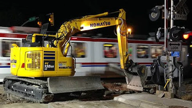 A yellow Komatsu excavator works at a grade crossing improvement site at night as a Utah Transit Authority light rail train passes in a blur of motion in the background. A railroad crossing signal is visible at right.