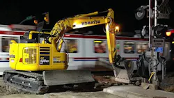 A yellow Komatsu excavator works at a grade crossing improvement site at night as a Utah Transit Authority light rail train passes in a blur of motion in the background. A railroad crossing signal is visible at right. A yellow Komatsu excavator works at a grade crossing improvement site at night as a Utah Transit Authority light rail train passes in a blur of motion in the background. A railroad crossing signal is visible at right.