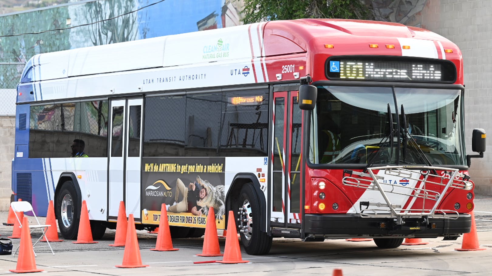 A Utah Transit Authority compressed natural gas bus, number 25001, navigates an orange traffic cone course during the APTA International Bus Roadeo competition. The red, white and blue bus displays a Markosian Auto advertisement on its side and shows Route 101 Marcus heading on its destination sign.