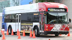 A Utah Transit Authority compressed natural gas bus, number 25001, navigates an orange traffic cone course during the APTA International Bus Roadeo competition. The red, white and blue bus displays a Markosian Auto advertisement on its side and shows Route 101 Marcus heading on its destination sign.