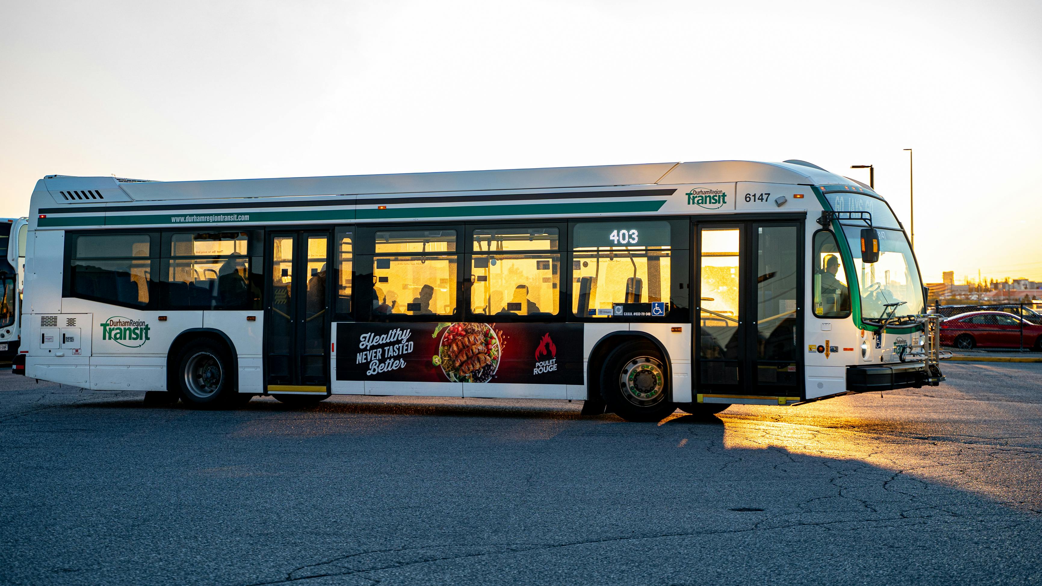 A Durham Region Transit bus, fleet number 6147, is photographed in a parking lot at golden hour. The white and green bus displays route number 403 and carries a side advertisement for Poulet Rouge restaurant. The bus faces right with its front door open, warm sunset light reflecting off the windows.