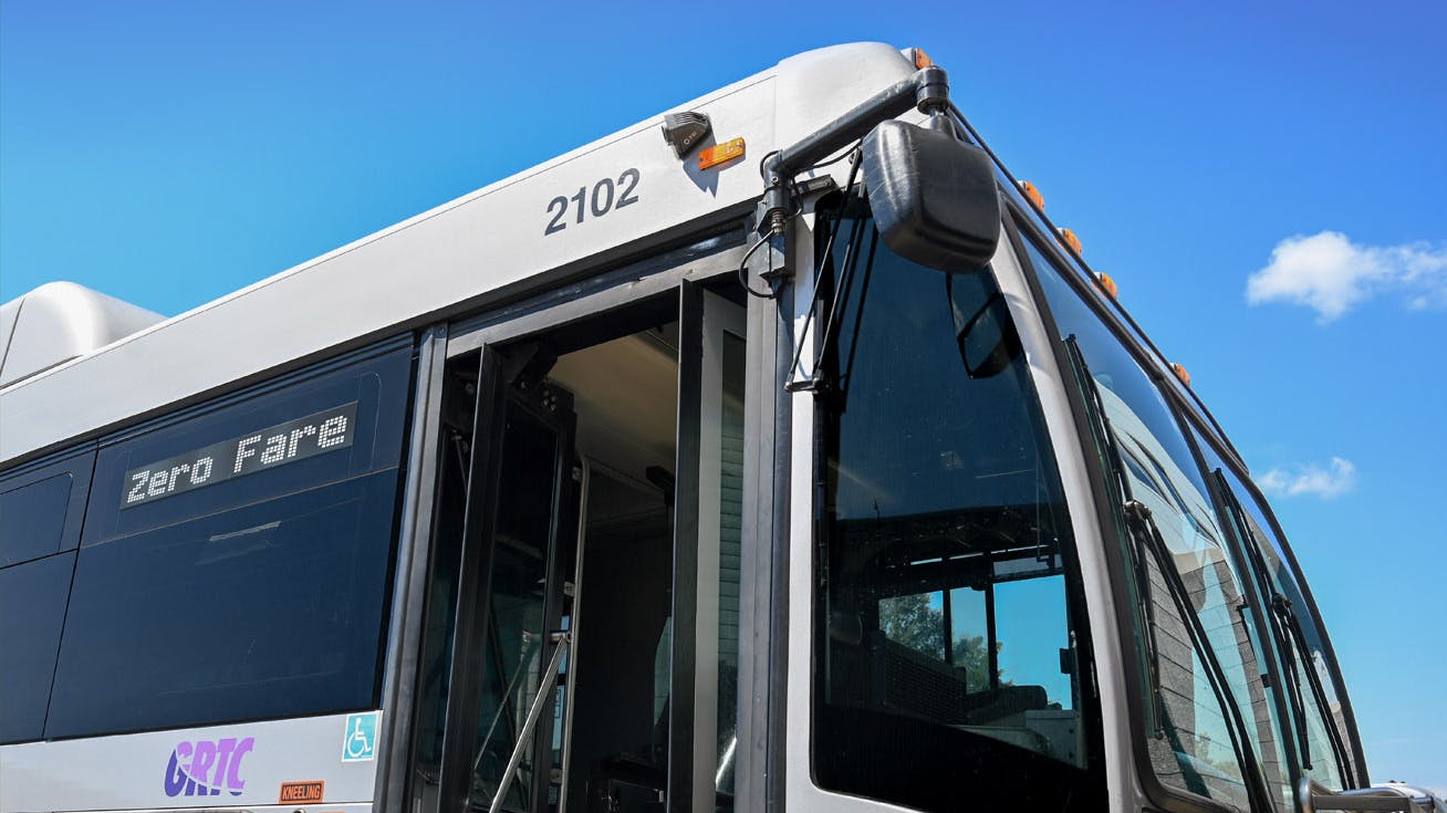 The front corner of a GRTC bus, fleet number 2102, photographed from a low angle against a bright blue sky with scattered clouds. The destination sign reads 'Zero Fare' and the front doors are open. The GRTC logo is visible in purple on the side panel near the accessibility symbol.