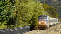 An MTA Metro-North Railroad locomotive, number 215, leads a passenger train along a wooded corridor in autumn. The blue and silver locomotive's headlights are illuminated as it approaches the camera, with a sound barrier wall running along the left side of the tracks and a tree-covered hillside in the background.
