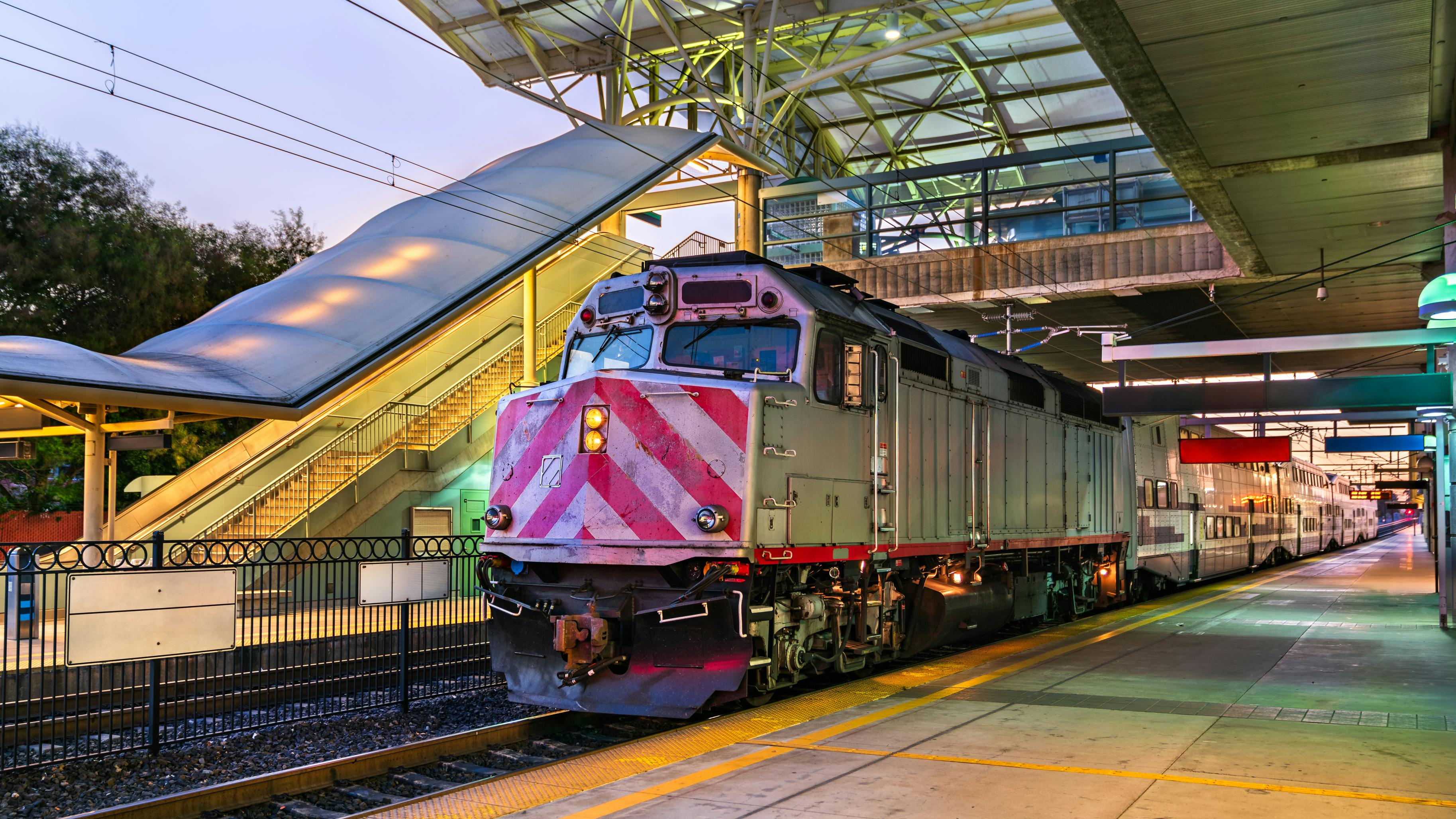 A commuter rail Caltrain locomotive with red and white chevron markings on its nose sits at a station platform at dusk, with double-deck passenger cars extending behind it. The station features a modern canopy structure with a glass and steel roof, an overhead pedestrian bridge, and a yellow safety stripe along the platform edge.
