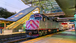 A commuter rail Caltrain locomotive with red and white chevron markings on its nose sits at a station platform at dusk, with double-deck passenger cars extending behind it. The station features a modern canopy structure with a glass and steel roof, an overhead pedestrian bridge, and a yellow safety stripe along the platform edge. A commuter rail Caltrain locomotive with red and white chevron markings on its nose sits at a station platform at dusk, with double-deck passenger cars extending behind it. The station features a modern canopy structure with a glass and steel roof, an overhead pedestrian bridge, and a yellow safety stripe along the platform edge.