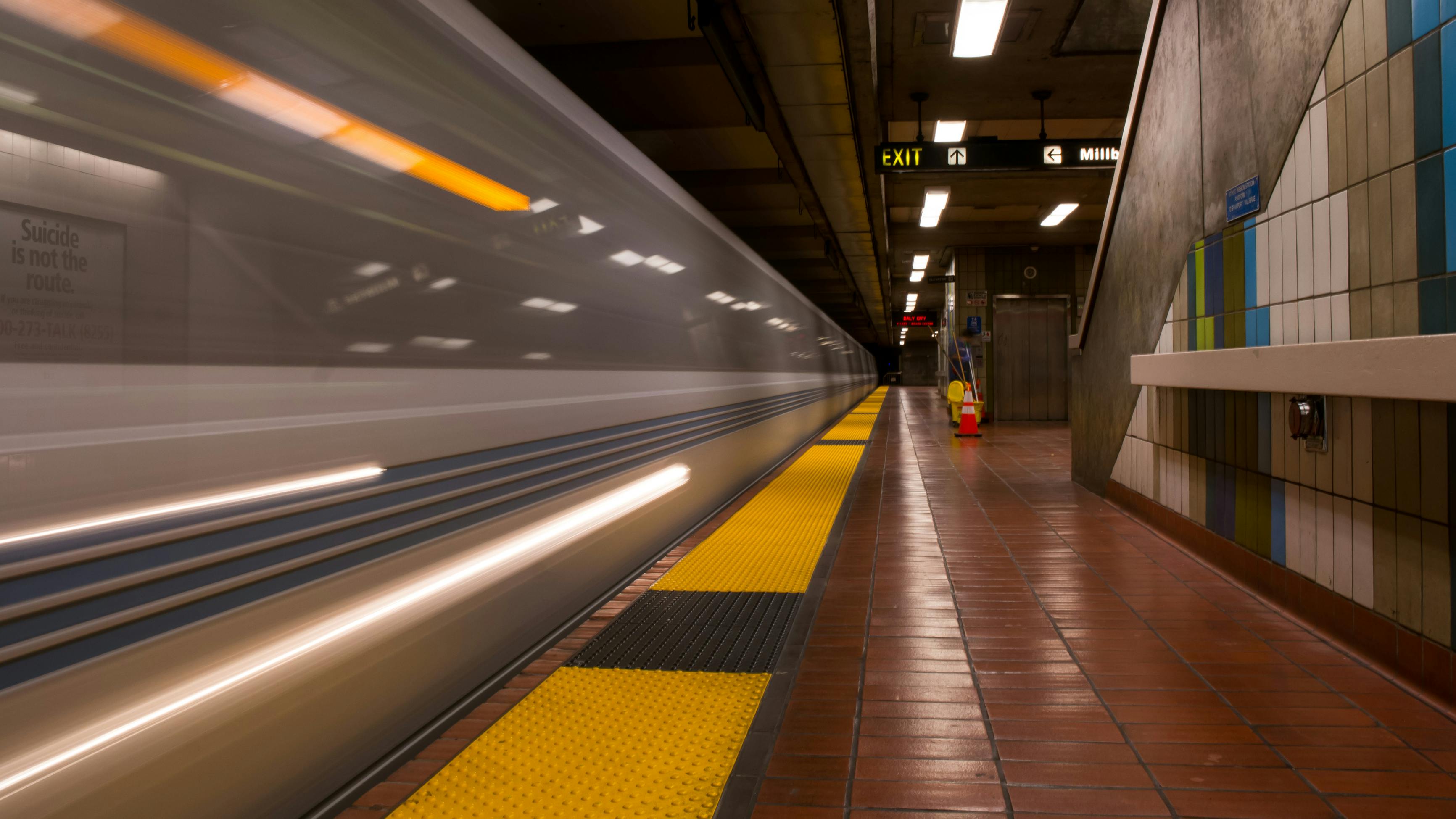 A BART train passes through an underground station with motion blur streaking along the platform edge. Overhead signage points to the exit and Millbrae, with a Daly City directional sign farther down the platform. The platform features a yellow tactile warning strip, red tile flooring and a geometric mosaic wall in blue, green and tan. A suicide prevention public service poster is partially visible at left.
