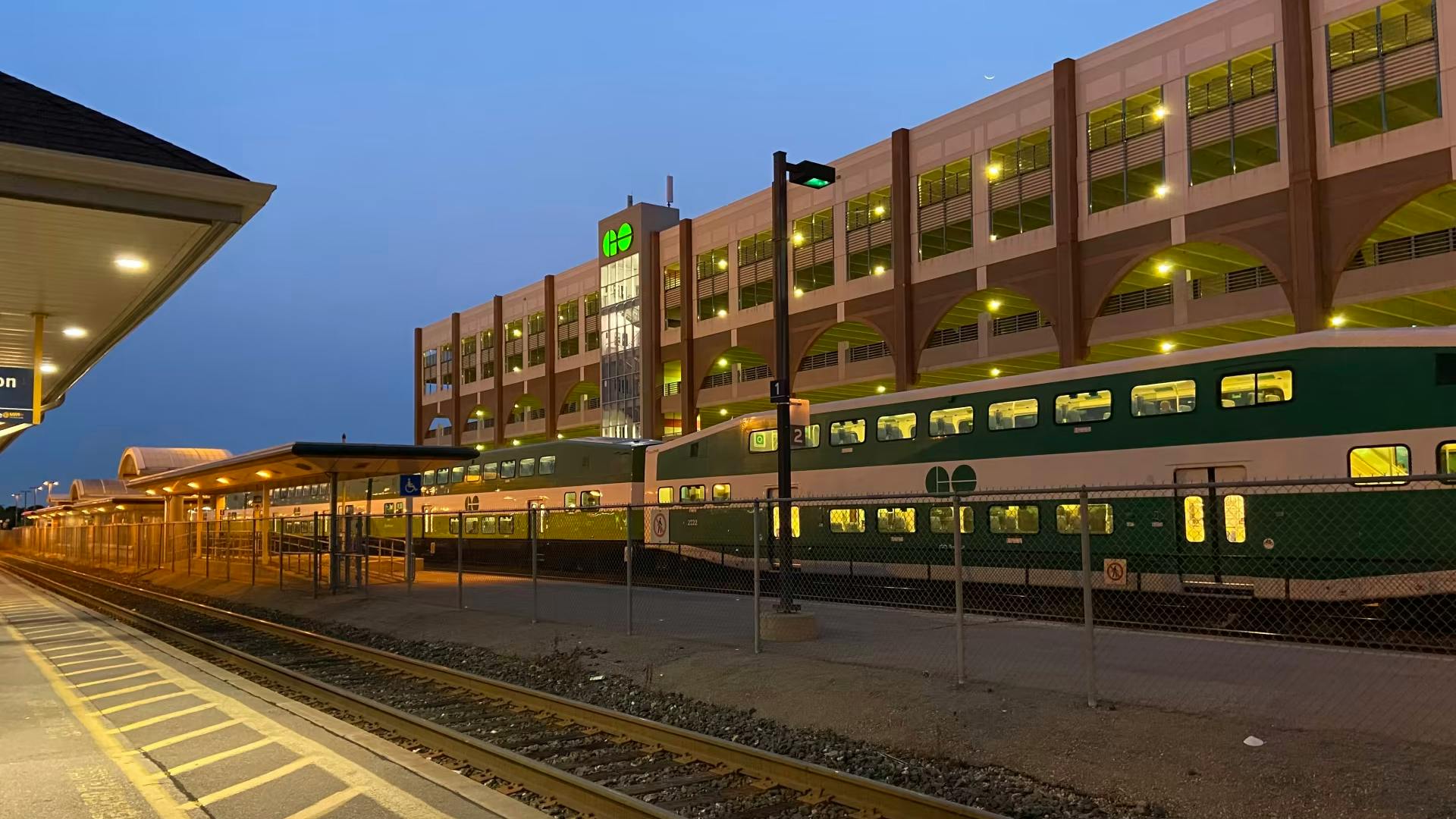 A GO Transit bi-level commuter train in green and white livery sits alongside a platform at dusk. A multi-story parking structure with arched openings and the GO Transit logo rises in the background, with platform canopies extending at left and tracks running through the foreground.