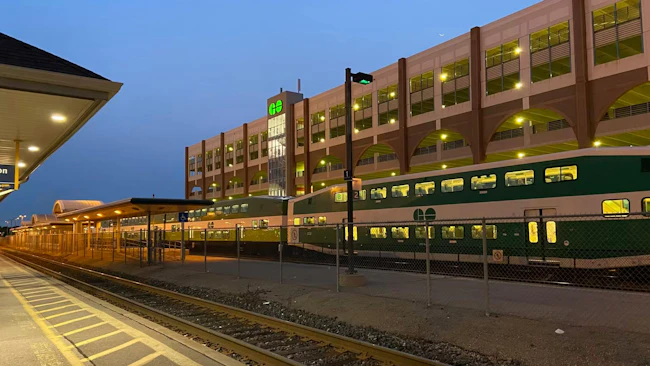 A GO Transit bi-level commuter train in green and white livery sits alongside a platform at dusk. A multi-story parking structure with arched openings and the GO Transit logo rises in the background, with platform canopies extending at left and tracks running through the foreground.