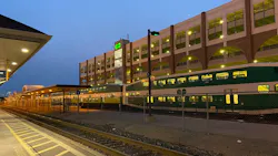 A GO Transit bi-level commuter train in green and white livery sits alongside a platform at dusk. A multi-story parking structure with arched openings and the GO Transit logo rises in the background, with platform canopies extending at left and tracks running through the foreground. A GO Transit bi-level commuter train in green and white livery sits alongside a platform at dusk. A multi-story parking structure with arched openings and the GO Transit logo rises in the background, with platform canopies extending at left and tracks running through the foreground.