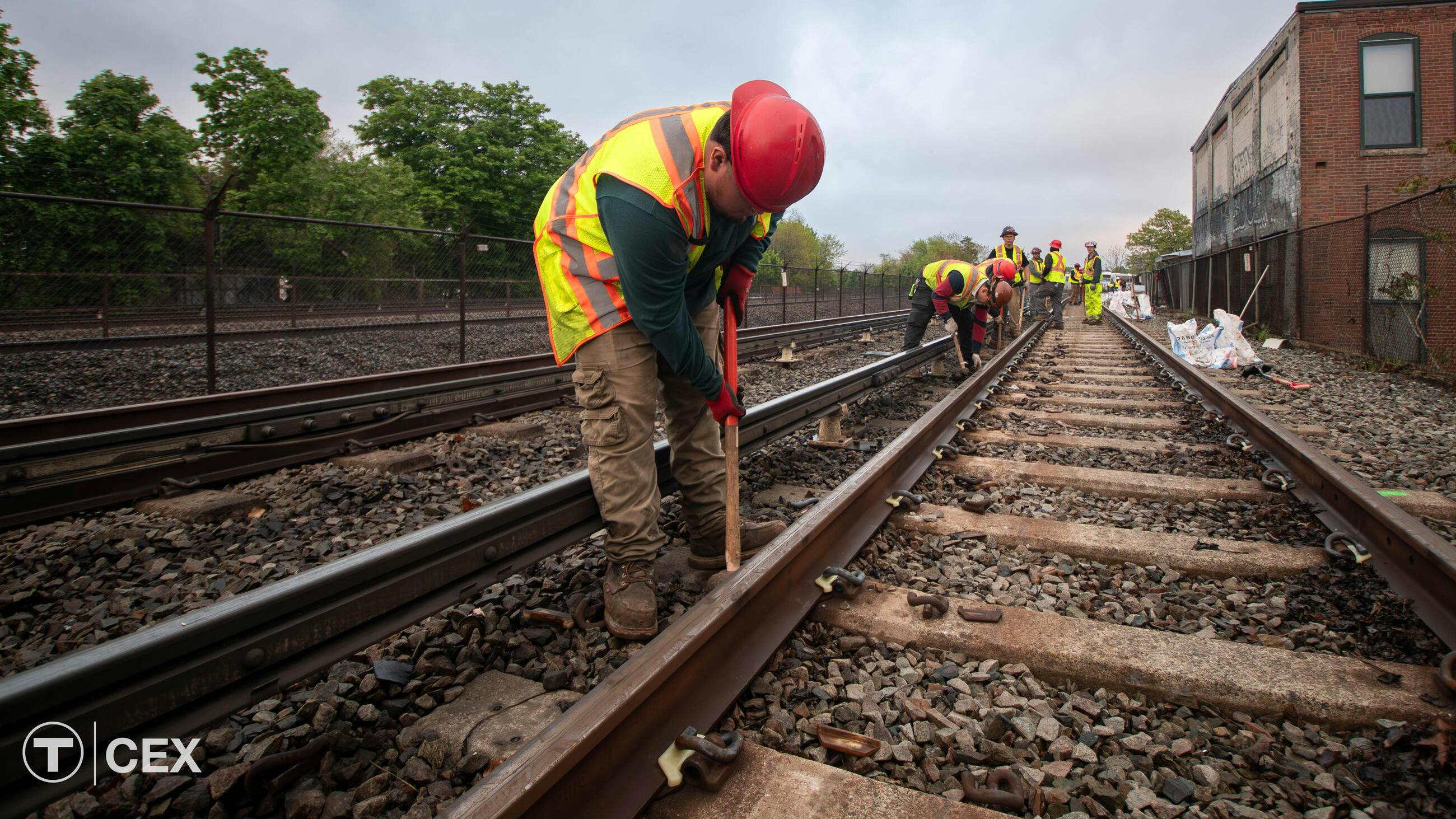A Massachusetts Bay Transportation Authority maintenance worker in a red hard hat and yellow safety vest bends to work on rail track ballast, with additional crew members working farther down the tracks and a brick building at right.