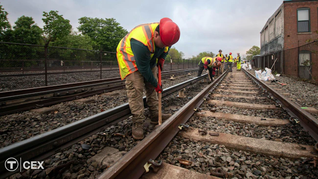 A Massachusetts Bay Transportation Authority maintenance worker in a red hard hat and yellow safety vest bends to work on rail track ballast, with additional crew members working farther down the tracks and a brick building at right.