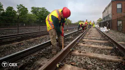 A Massachusetts Bay Transportation Authority maintenance worker in a red hard hat and yellow safety vest bends to work on rail track ballast, with additional crew members working farther down the tracks and a brick building at right. A Massachusetts Bay Transportation Authority maintenance worker in a red hard hat and yellow safety vest bends to work on rail track ballast, with additional crew members working farther down the tracks and a brick building at right.