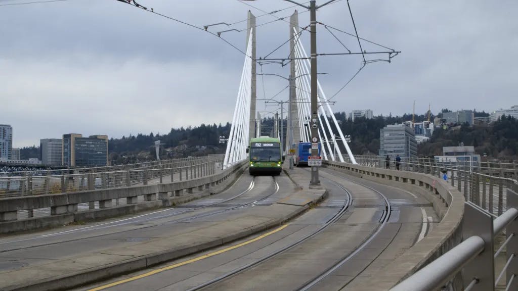 A green TriMet FX articulated bus crosses the cable-stayed Tilikum Crossing bridge in Portland, Oregon, sharing the transit-only deck with light rail tracks, with the city skyline and overcast sky in the background.
