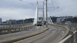 A green TriMet FX articulated bus crosses the cable-stayed Tilikum Crossing bridge in Portland, Oregon, sharing the transit-only deck with light rail tracks, with the city skyline and overcast sky in the background.