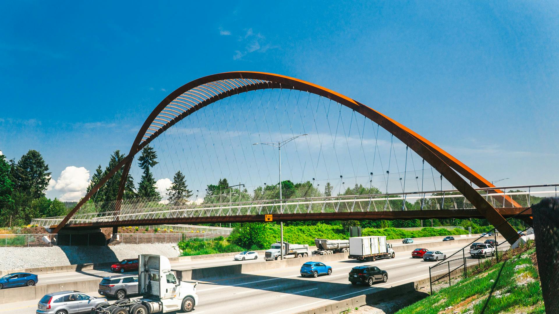 A new steel arch pedestrian and transit overpass with a Corten-style rust-colored finish spans a multi-lane highway, with cars, trucks and a tractor-trailer passing beneath on a clear day.