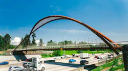 A new steel arch pedestrian and transit overpass with a Corten-style rust-colored finish spans a multi-lane highway, with cars, trucks and a tractor-trailer passing beneath on a clear day.