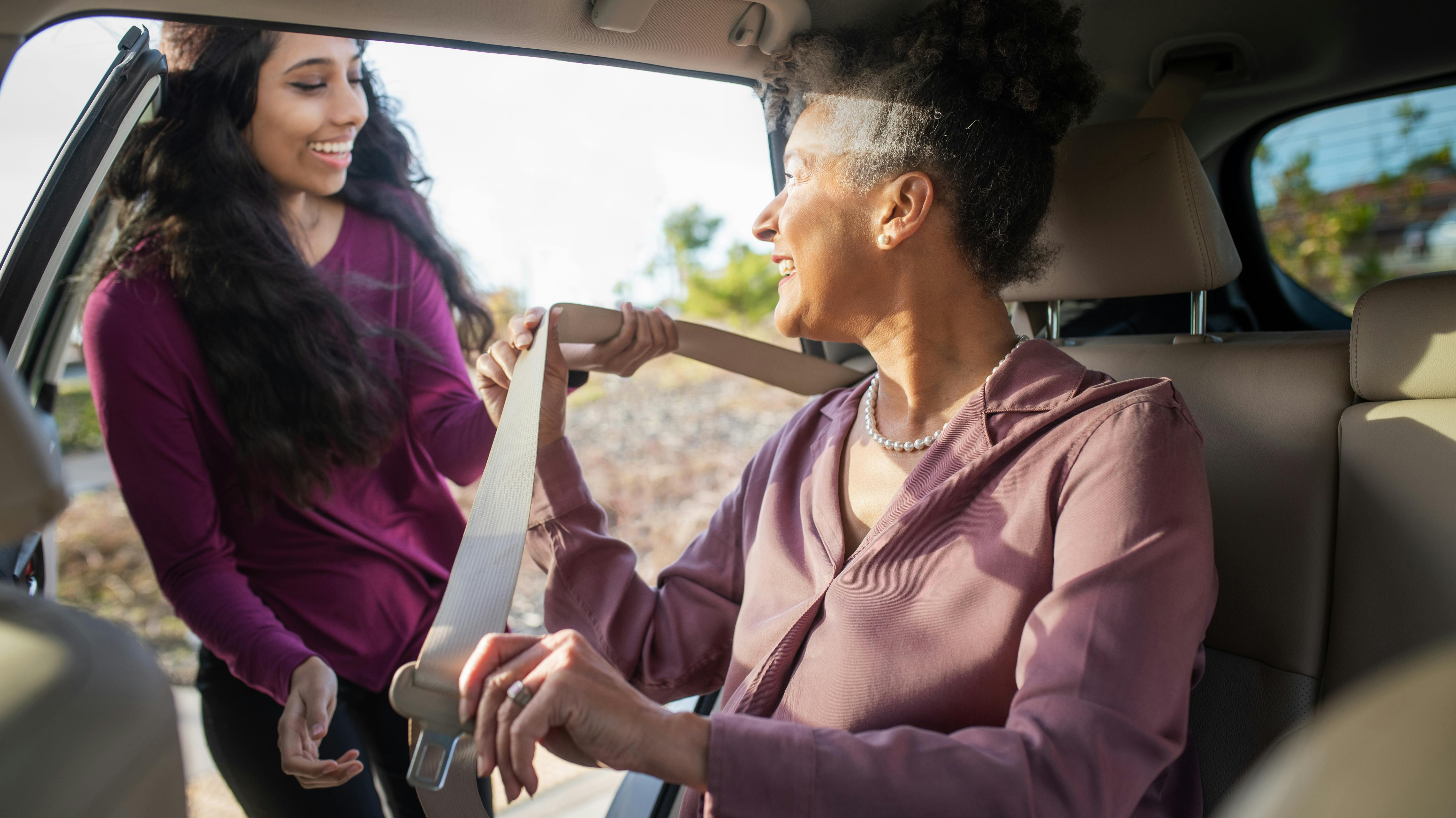 A younger woman in a maroon top smiles as she helps an older woman in a dusty pink blouse buckle her seatbelt in the back seat of a car, with sunlight streaming through the open door.