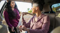 A younger woman in a maroon top smiles as she helps an older woman in a dusty pink blouse buckle her seatbelt in the back seat of a car, with sunlight streaming through the open door.