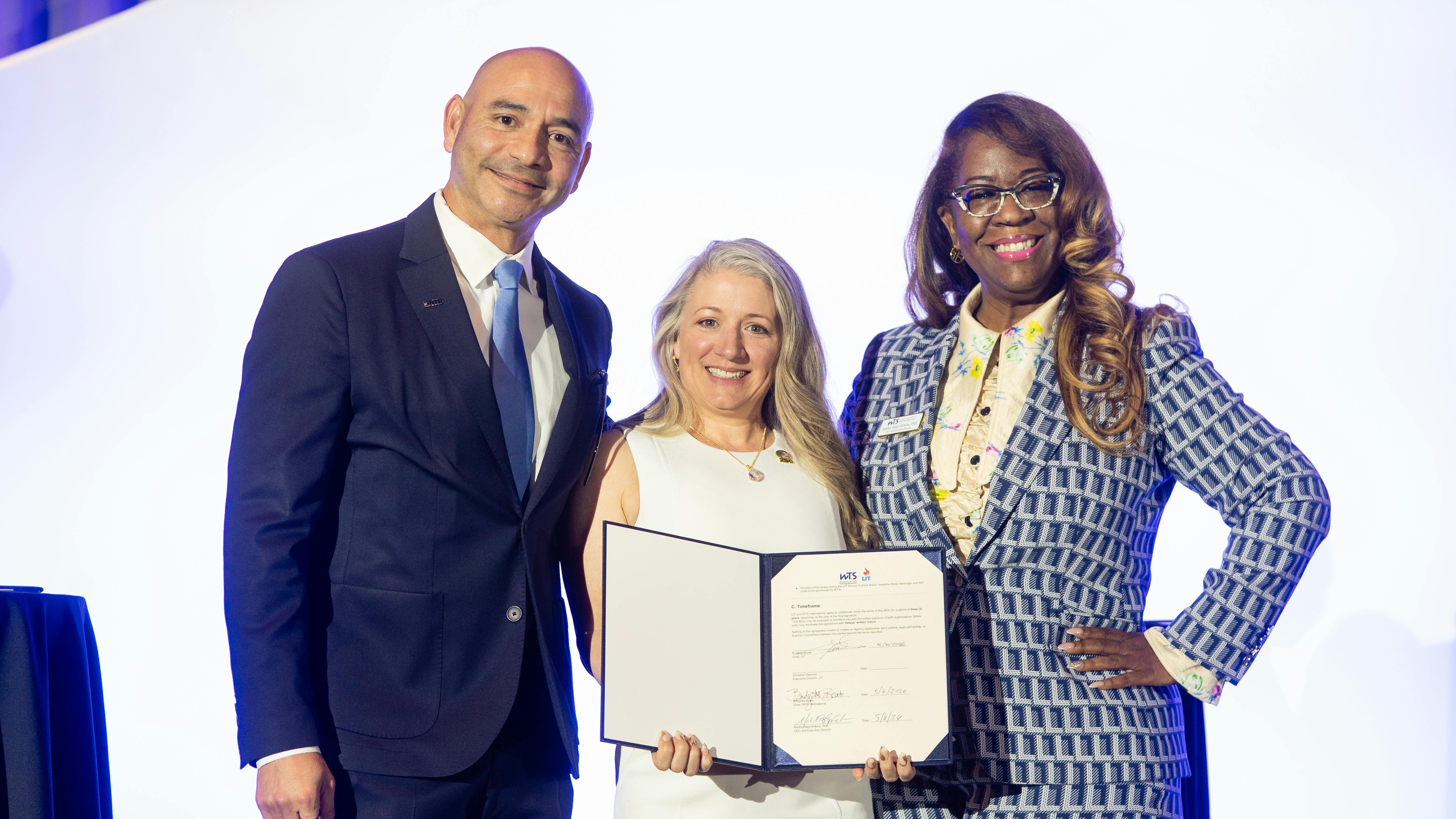 LIT Chair Eugenio Bonet, WTS International Chair Bridgette Beato and WTS International CEO Malika Reed Wilkins pose on stage holding an open signed memorandum of understanding between the two organizations.