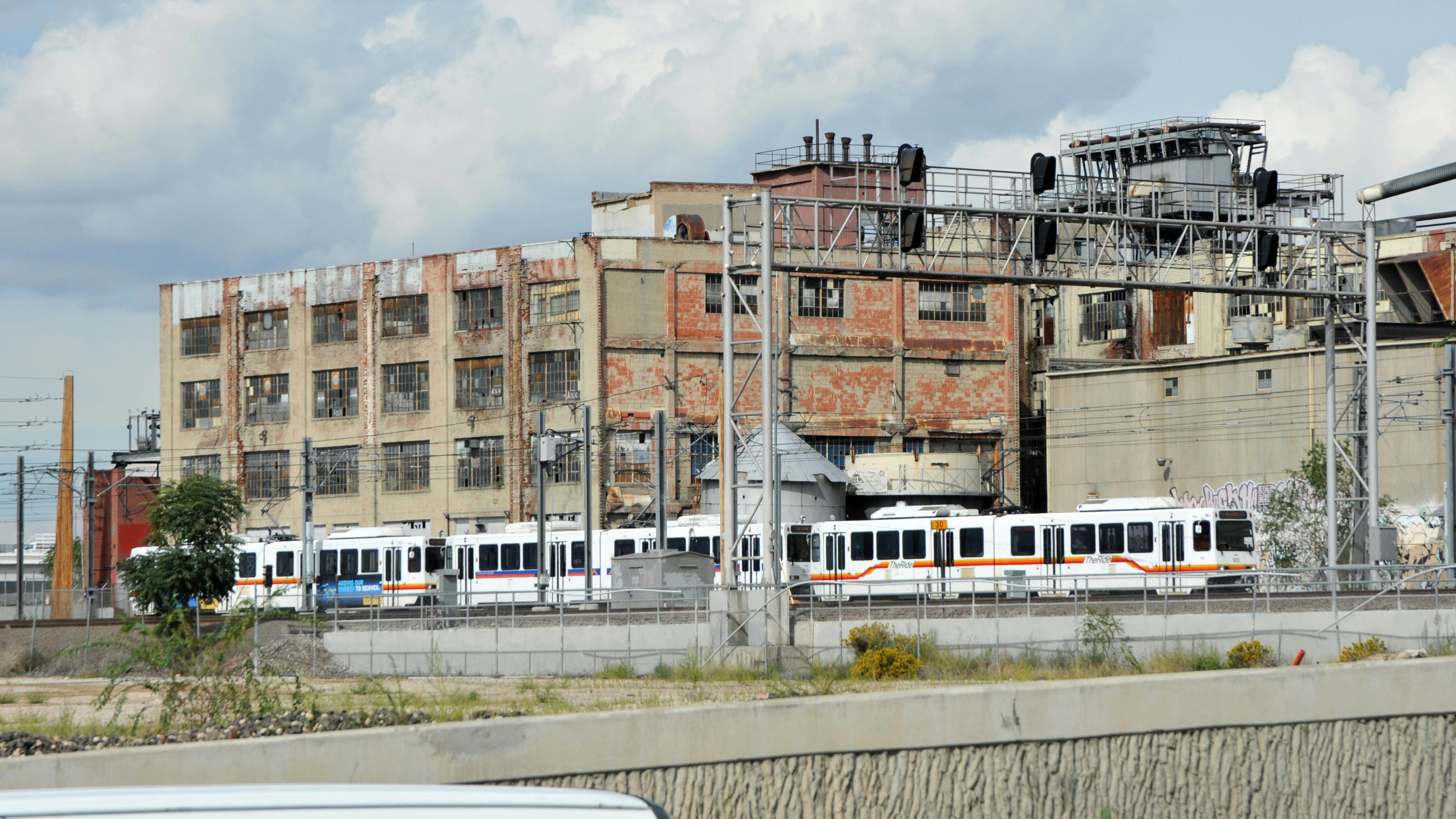 A two-car RTD light-rail train in white with red, yellow and blue striping passes a weathered multi-story industrial building with peeling brick and broken windows under a partly cloudy sky.