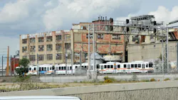 A two-car RTD light-rail train in white with red, yellow and blue striping passes a weathered multi-story industrial building with peeling brick and broken windows under a partly cloudy sky. A two-car RTD light-rail train in white with red, yellow and blue striping passes a weathered multi-story industrial building with peeling brick and broken windows under a partly cloudy sky.