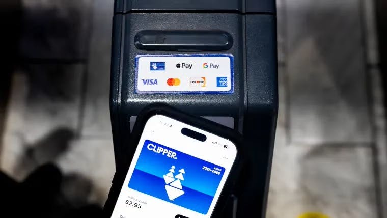 A smartphone displaying a digital Clipper transit card with a $2.95 balance is held up to a contactless fare reader showing Apple Pay, Google Pay, Visa, Mastercard, Discover and American Express acceptance logos.