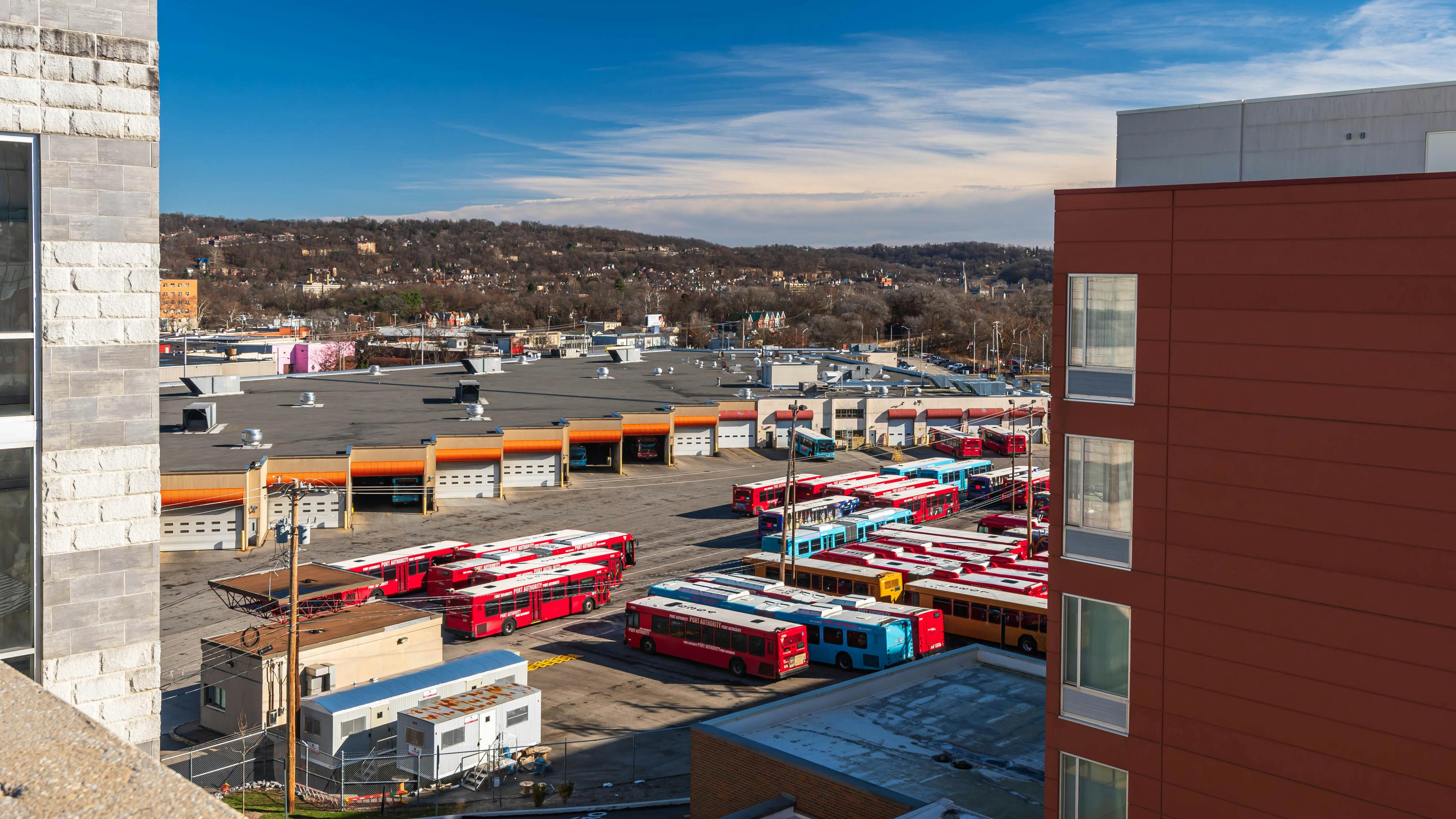 An overhead view of a Pittsburgh Regional Transit bus garage and yard, with rows of red, blue and yellow Port Authority-branded buses parked outside maintenance bays, framed by surrounding buildings and tree-covered hillsides in the distance.