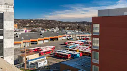 An overhead view of a Pittsburgh Regional Transit bus garage and yard, with rows of red, blue and yellow Port Authority-branded buses parked outside maintenance bays, framed by surrounding buildings and tree-covered hillsides in the distance. An overhead view of a Pittsburgh Regional Transit bus garage and yard, with rows of red, blue and yellow Port Authority-branded buses parked outside maintenance bays, framed by surrounding buildings and tree-covered hillsides in the distance.