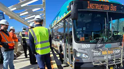 Denver RTD staff in hard hats and high-visibility vests gather alongside a teal and silver New Flyer Free MetroRide bus marked 'Instruction Bus' as it pulls up to a platform along the East Colfax Bus Rapid Transit corridor in Denver.